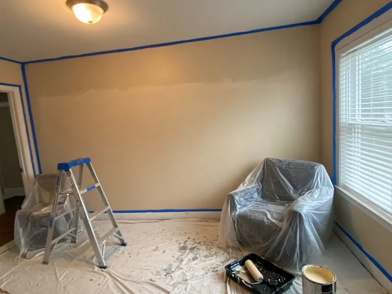 Room prepared for painting with blue tape, drop cloth, covered chair, ladder and paint tray